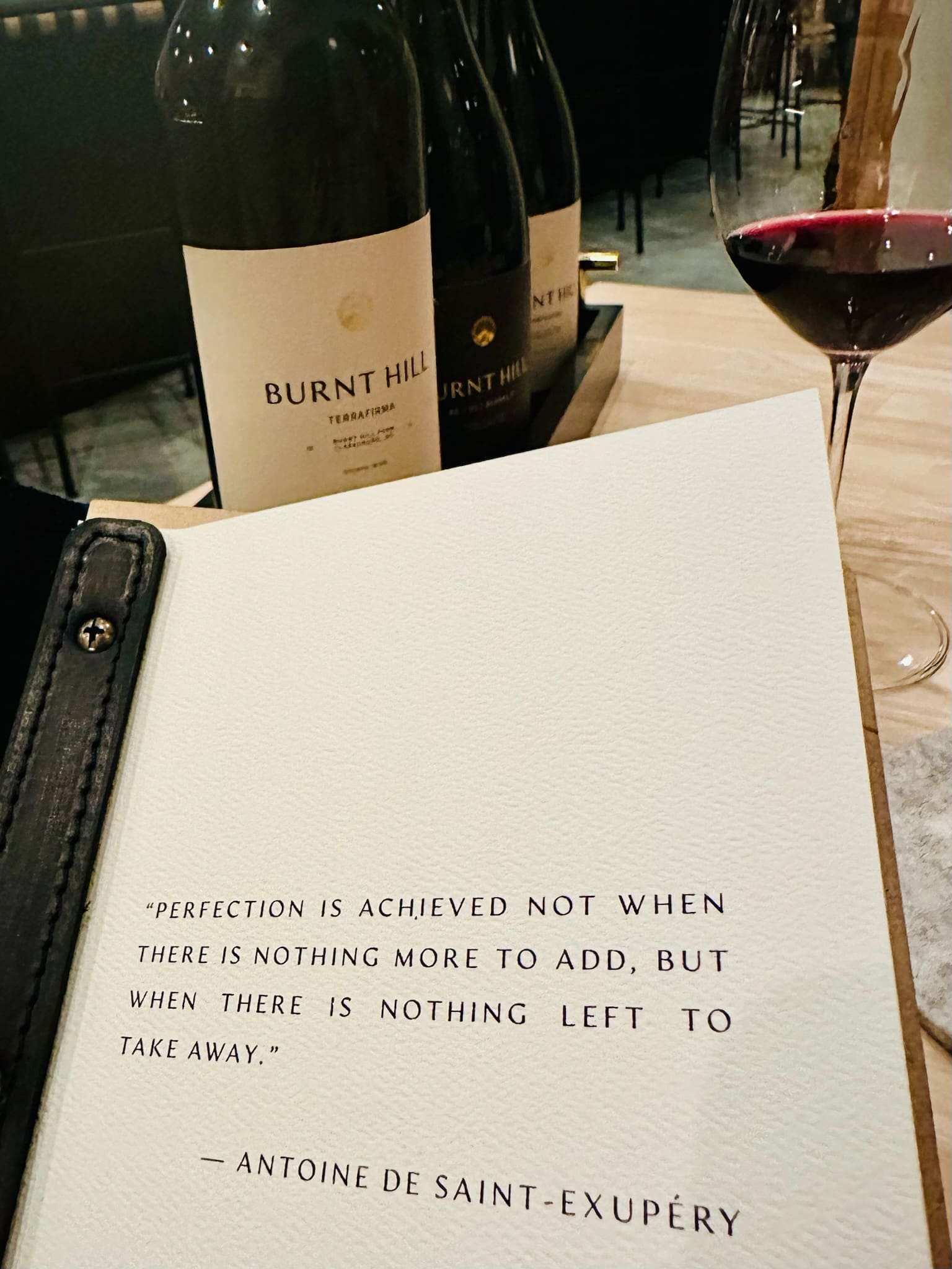 a Close up of an Open Book with a Quote by Antoine De Saint exupéry Beside a Glass of Red Wine and Three Burnt Hill Farm Bottles on a Wooden Table in a Dimly Lit Elegant Setting