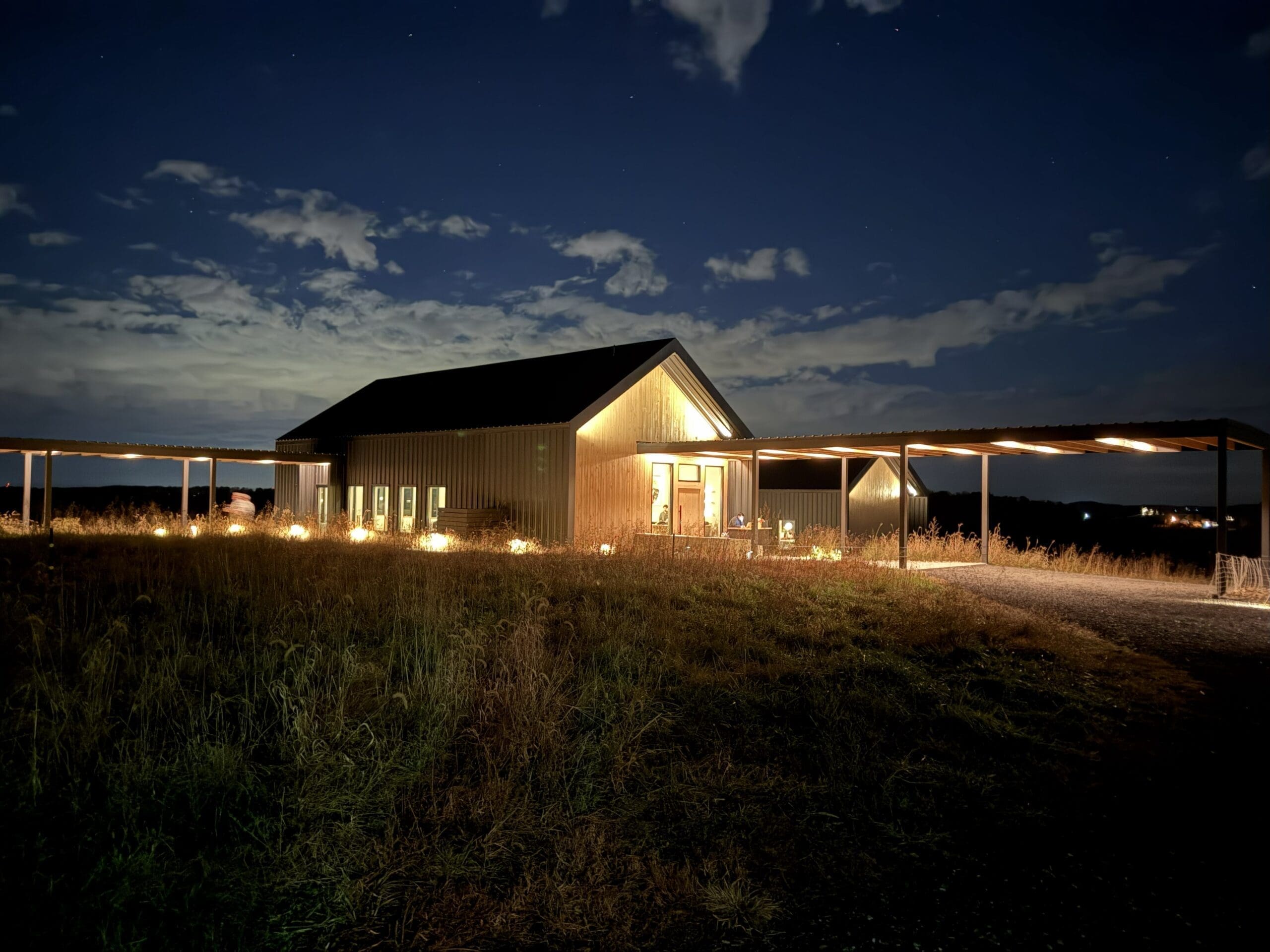 a Warmly Lit Barn Style Building Stands Alone at Night Under a Partly Cloudy Sky in Mt Airy Md Soft Lights Illuminate Windows and a Covered Walkway While Tall Grass Surrounds the Structure Creating a Peaceful Rural Scene