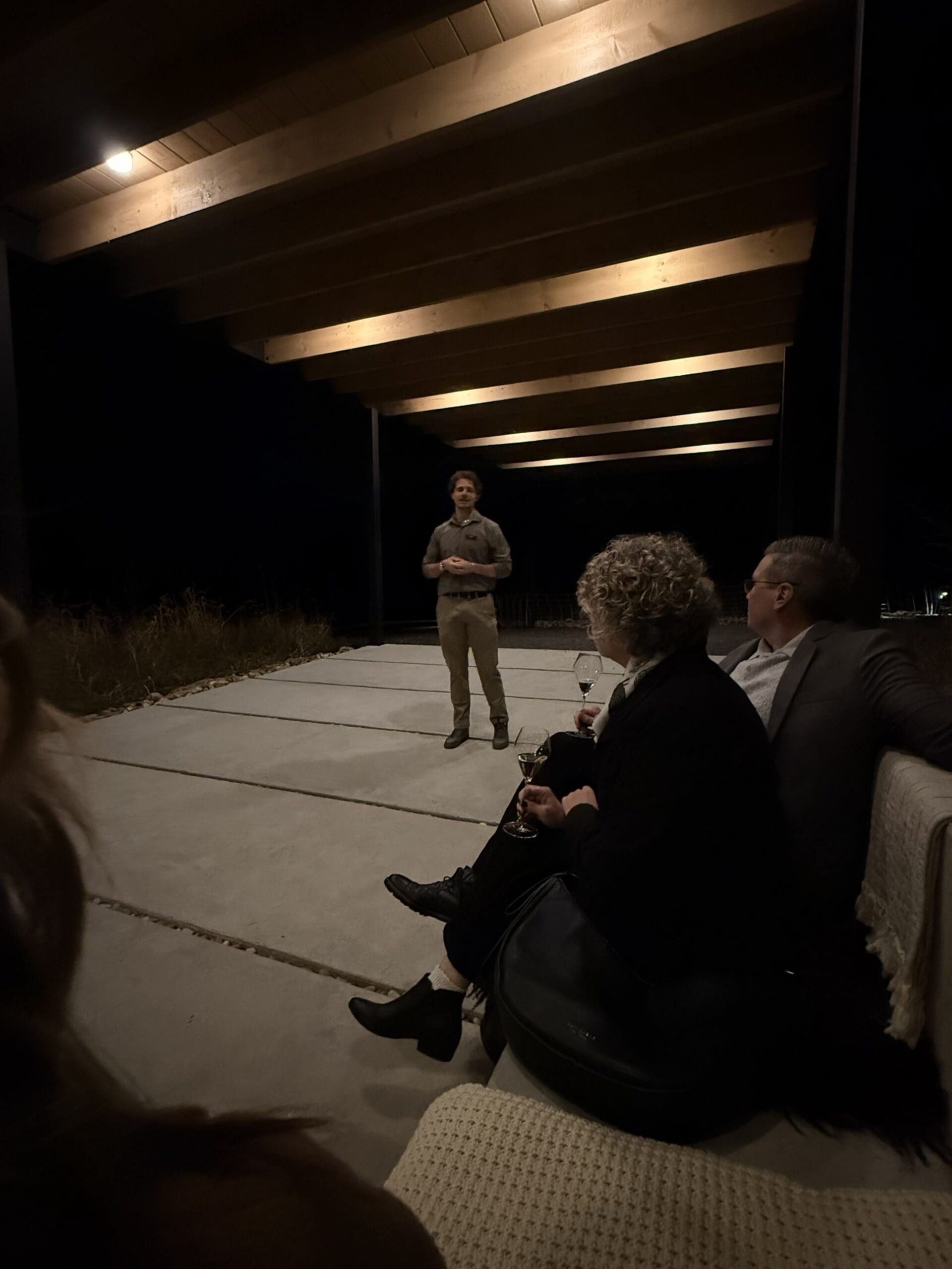 a Man Stands Speaking Under Wooden Ceiling Beams at Night at Burnt Hill Farm Facing Three Seated People Two Sit on a Cushioned Outdoor Sofa Holding Drinks While the Third is Partly Visible the Scene is Dimly Lit by Warm Overhead Lights