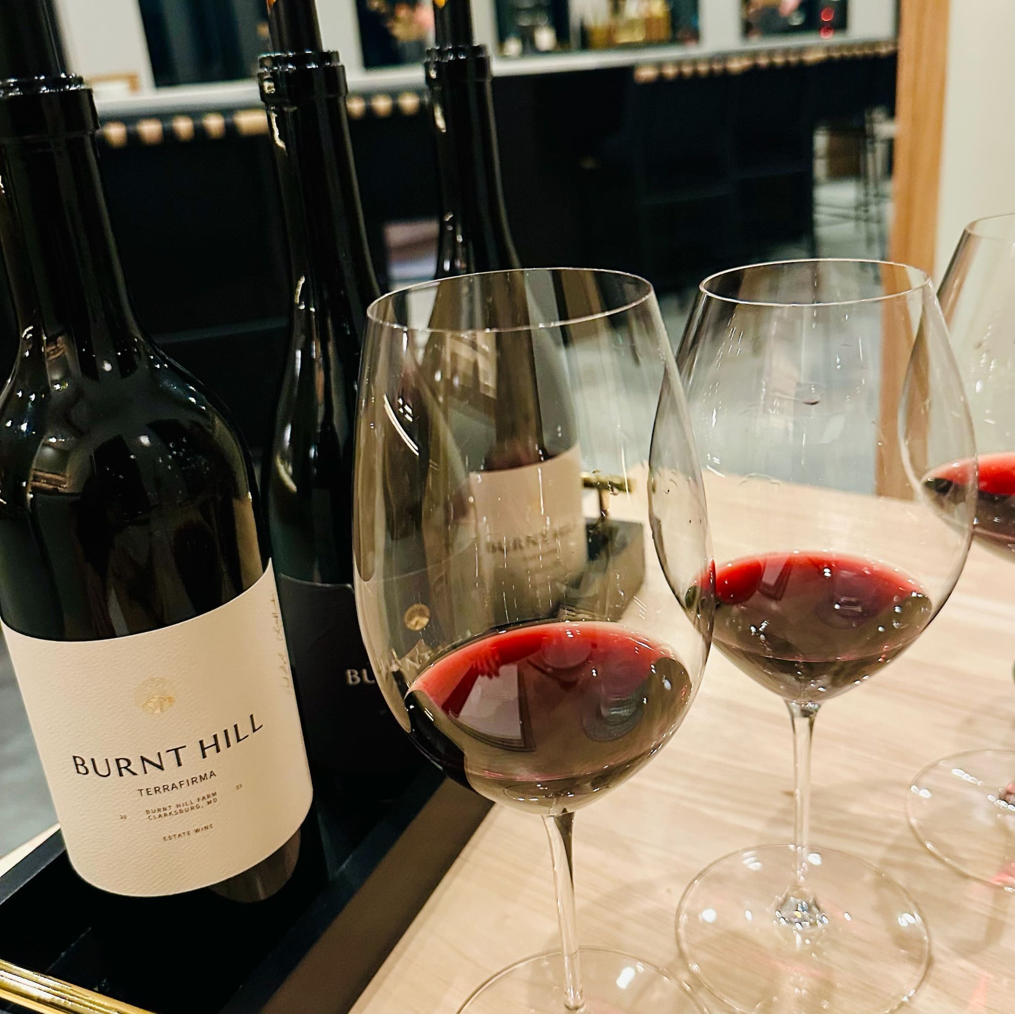 Three Wine Glasses Filled with Red Wine Are Arranged Before Three Burnt Hill Farm Bottles on a Wooden Table a Sleek Modern Bar with Reflective Shelves Forms the Backdrop Setting the Scene for an Elegant Wine Tasting Event