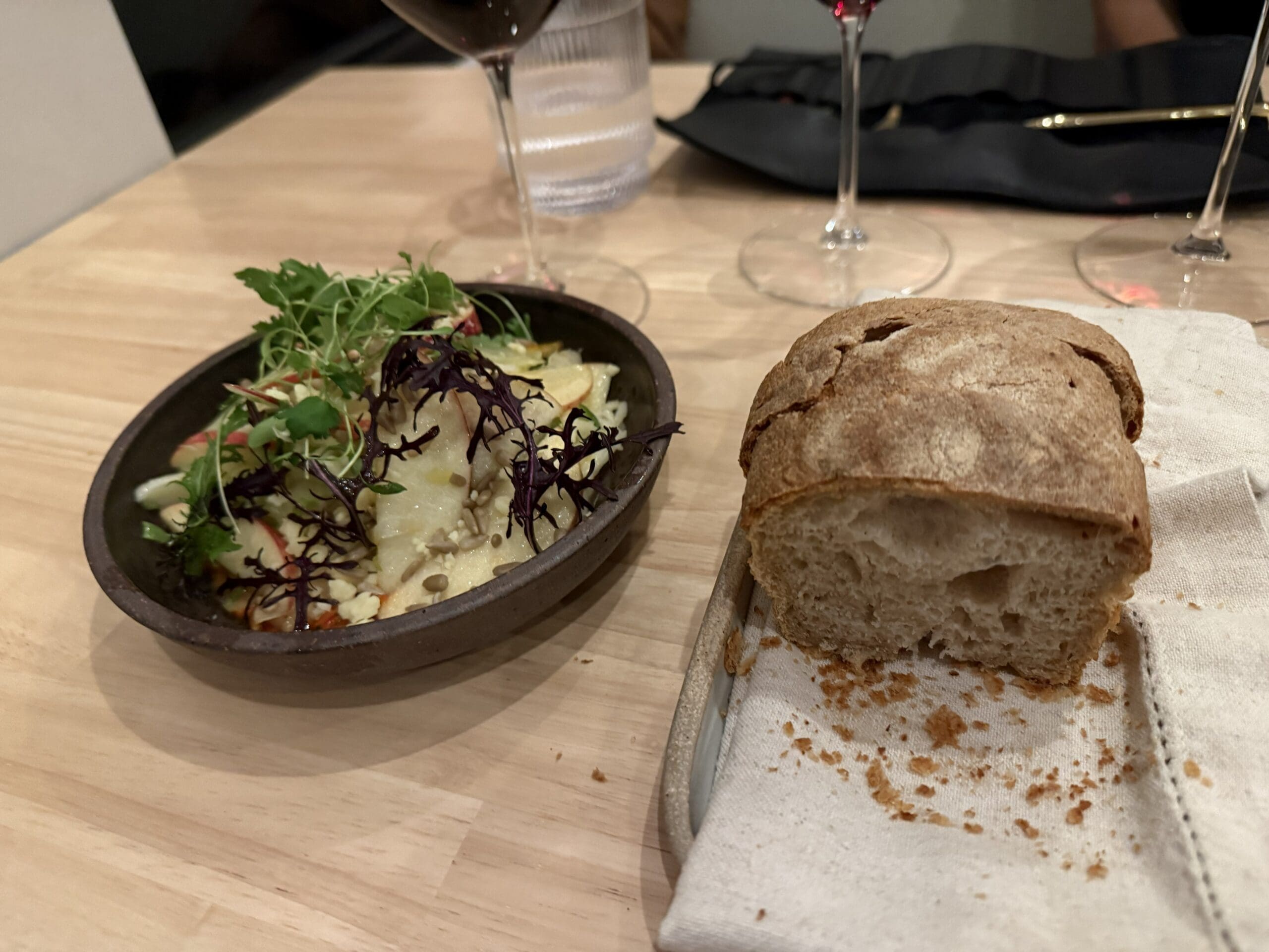 a Slice of Rustic Burnt Hill Farm Bread on a Cloth napkin lined Plate Sits Beside a Bowl of Salad Topped with Microgreens Two Glasses of Red Wine and a Clear Glass of Water Are in the Background on a Light Wooden Table