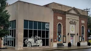 a Two Story Brick and Concrete Building in Mt Airy Md with Large Windows on the Left Side Displaying a Mural of a Vintage Car the Right Side Features Classical Architectural Details a Triangle Pediment a Clock and Arched Windows a Bench Sits Outside the Entrance