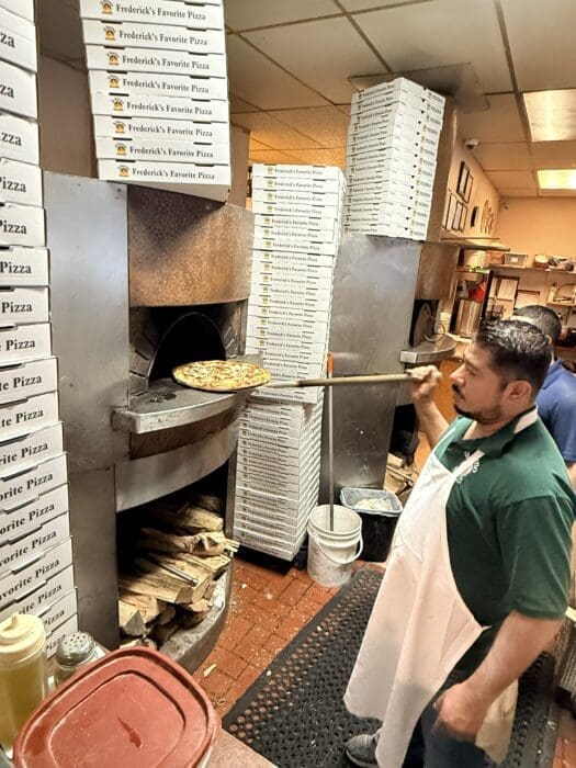 a Man in a Green Shirt and White Apron Uses a Long Pizza Peel to Remove Pizza in Frederick Md from a Wood Fired Oven Stacks of Fredericks Favorite Pizza Boxes Surround Him with Firewood Stored Below the Oven Inside a Busy Pizzeria Kitchen