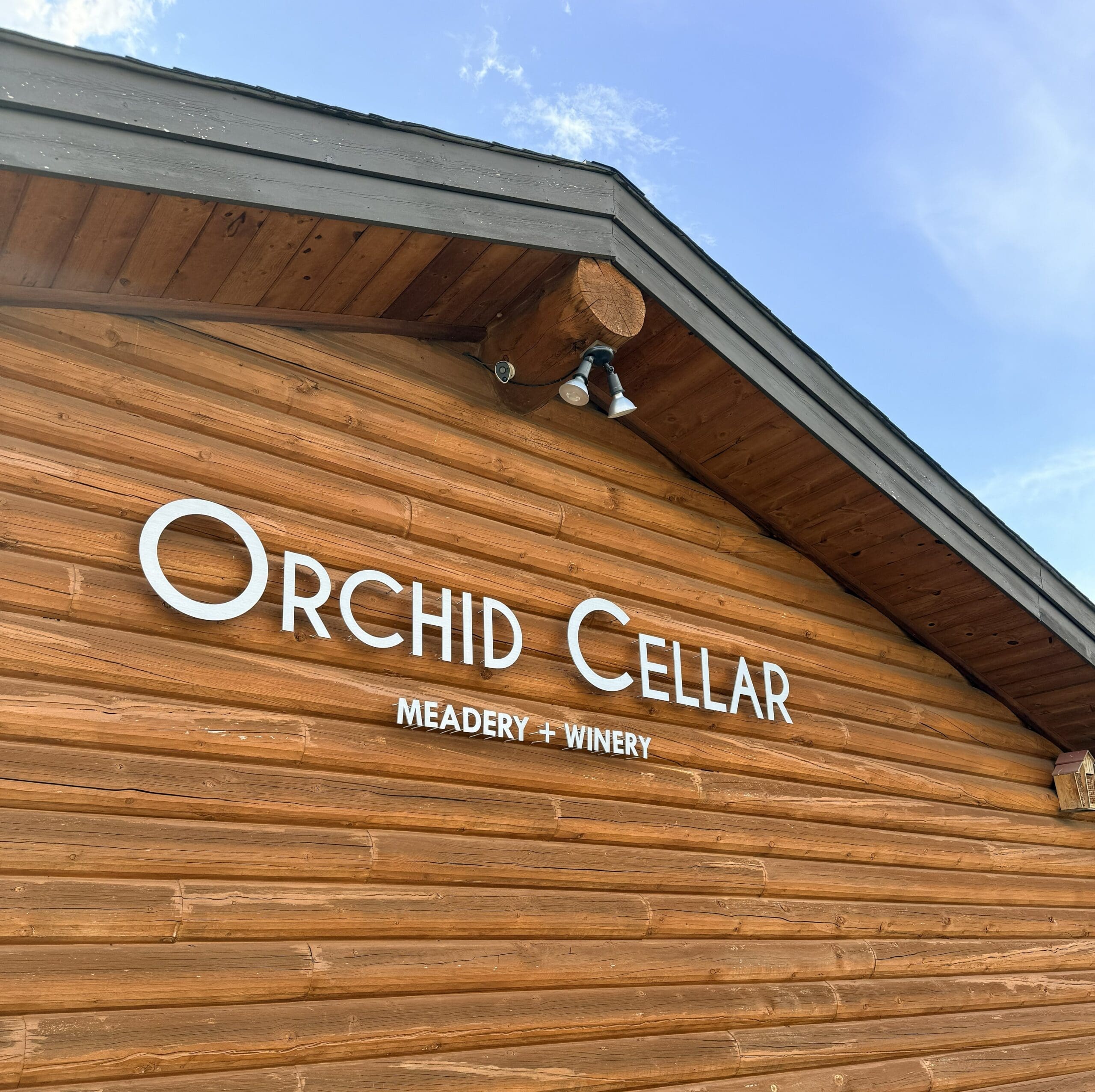 the Image Shows a Wooden Building with a Sloped Roof Against a Blue Sky in Frederick Maryland on the Wood Siding Large White Letters Read Orchid Cellar and Below in Smaller Text Meadery + Winery Two Outdoor Lights Are Mounted Above the Sign