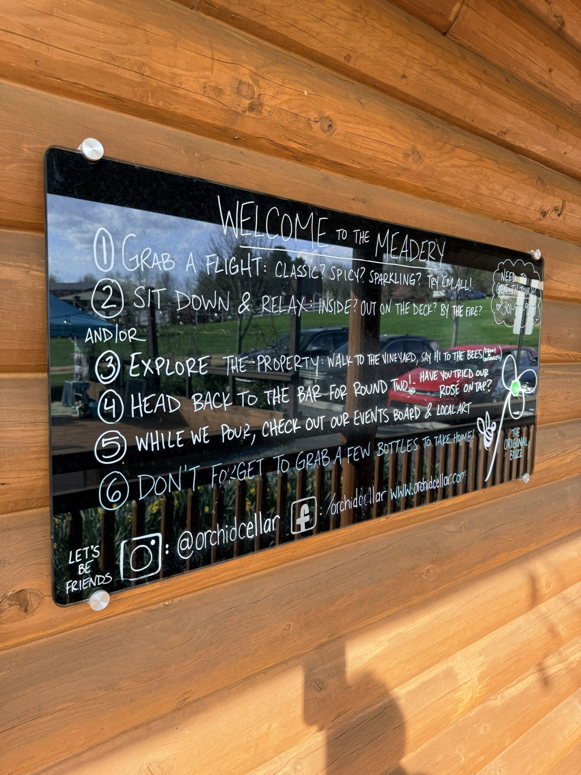 a Glossy Black Sign on a Wooden Wall at Orchid Cellar Middletown Md Reads Welcome to the Meadery and Lists Five Steps for Visiting from Trying Mead Flights to Grabbing Bottles Social Media Handles and Playful Doodles Decorate the Sign