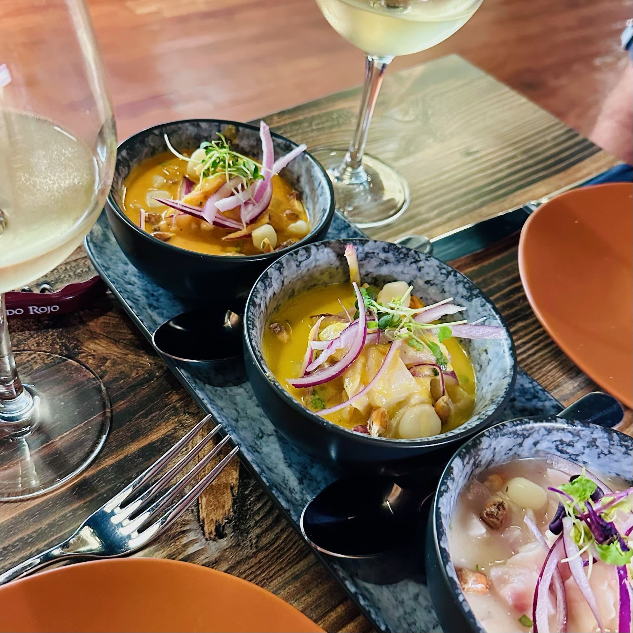 Three Bowls of Orange Soup Topped with Red Onion Slices and Microgreens on a Rustic Wooden Table with Wine Glasses Nearby