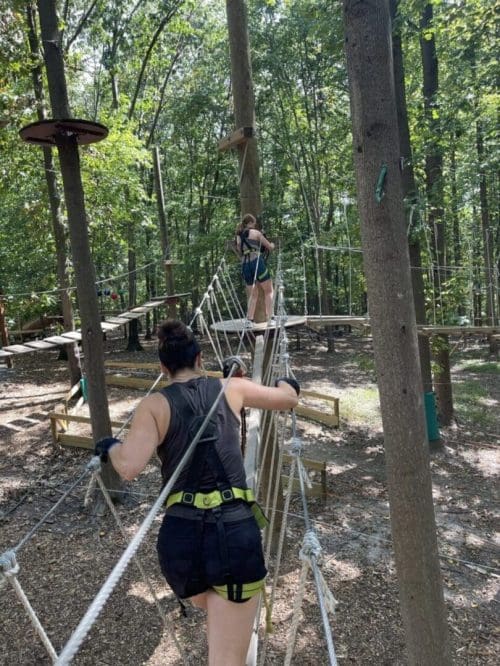 Two people wearing harnesses and gloves cross a rope bridge in a forested adventure park near New Market MD. Sunlight filters through the trees onto the woodchip-covered ground as one person grips the ropes, while the other moves confidently ahead.
