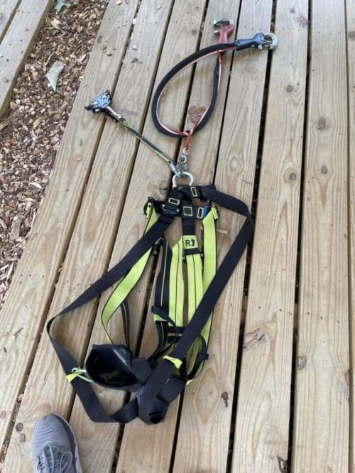 A yellow and black safety harness with attached lanyard and metal clips is laid out on a wooden deck in New Market, MD. The harness is loosened and unbuckled, spread out flat. A person’s gray shoe is visible at the lower left corner of the image.