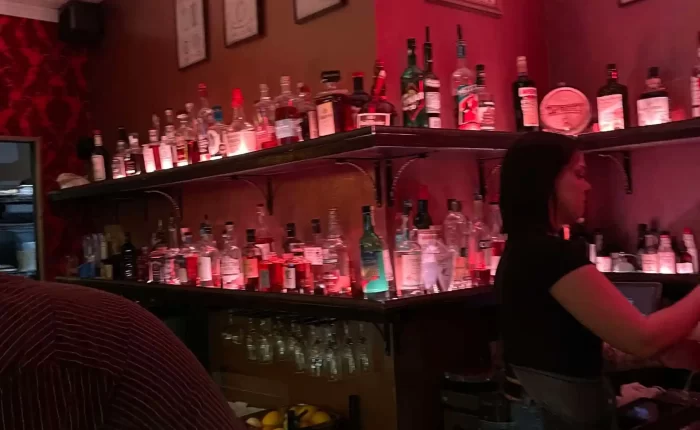 A dimly lit bar with red walls, shelves stocked with bottles of liquor, and framed art—just the kind of cozy spot you’d find in Bend, Oregon. A bartender prepares drinks as a person in a striped shirt sits at the bar in the foreground.